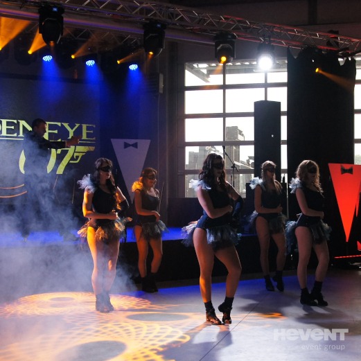 Grupa tancerek w maskach i kostiumach w stylu Jamesa Bonda na scenie z oświetleniem eventowym i logiem GoldenEye 007 w tle, podczas występu z wytwornicą dymu.