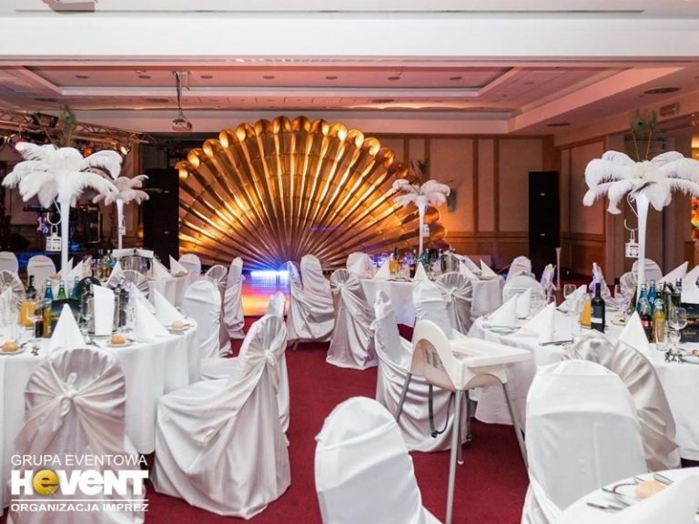 Uroczysta sala bankietowa z okrągłymi stołami nakrytymi białymi obrusami, krzesła z białymi pokrowcami, dekoracje z piór, złota dekoracja w kształcie muszli na tle, czerwona wykładzina.