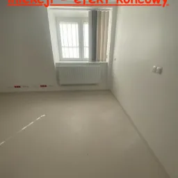 Jasne pomieszczenie piwniczne po pracach zabezpieczających, z oknem z kratami i grzejnikiem, świeżo pomalowane ściany i podłoga z widocznymi gniazdkami elektrycznymi.