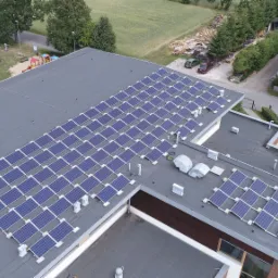 Widok z góry na dużą instalację paneli słonecznych na płaskim dachu budynku, z widocznym placem zabaw i zaparkowanymi samochodami w tle.