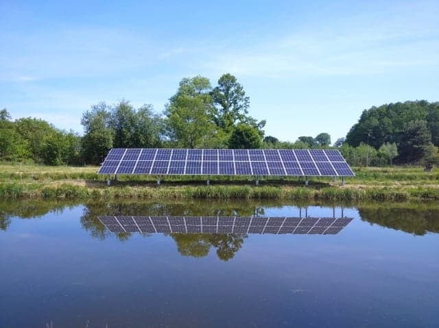 Instalacja paneli słonecznych nad stawem, z wyraźnym odbiciem w wodzie i zielonymi drzewami w tle, w słoneczny dzień.