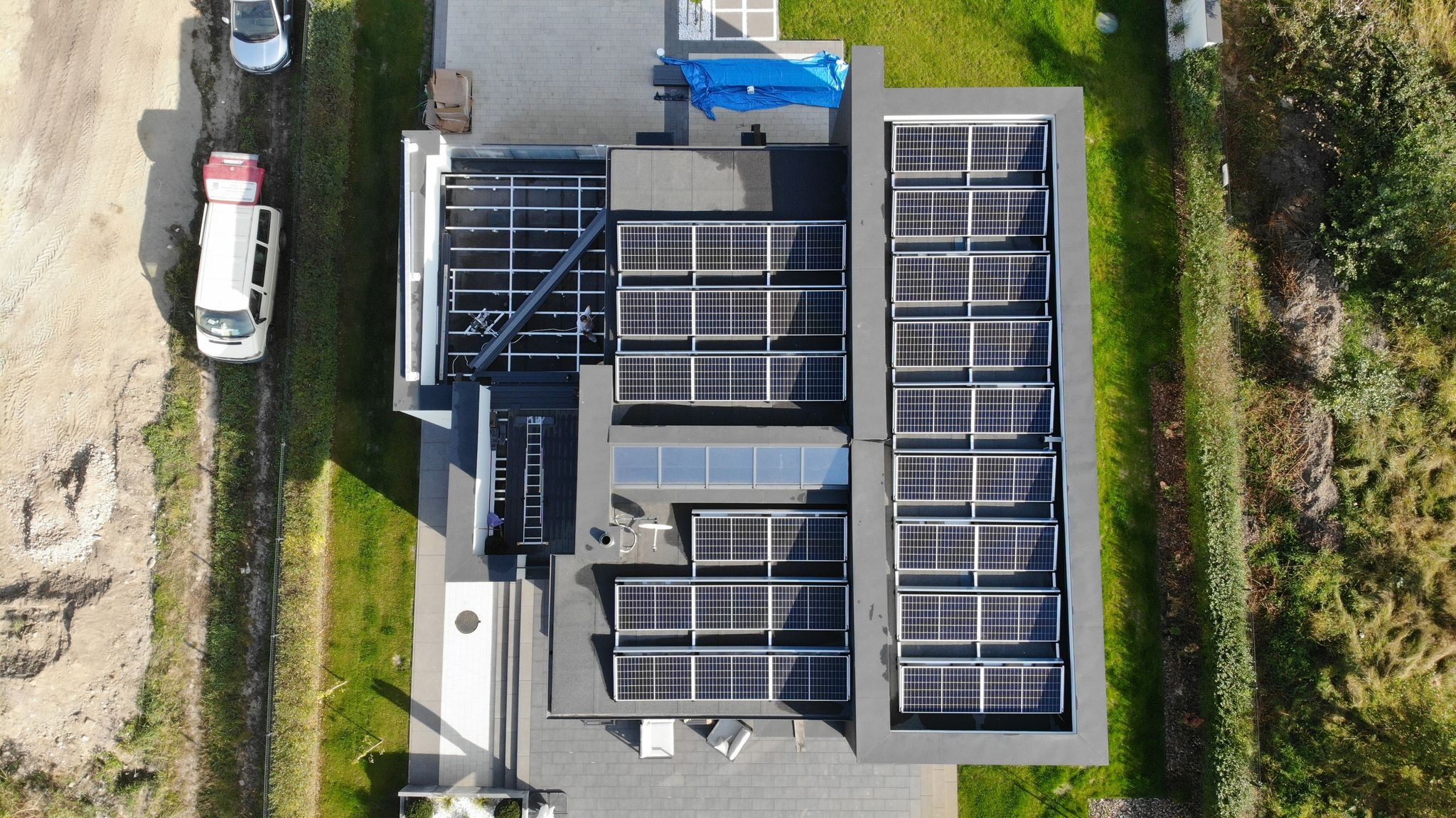 Widok z góry na dach budynku z częściowo zamontowanymi panelami słonecznymi, widoczna drabina i konstrukcja wsporcza, obok zaparkowane samochody i zielony trawnik.