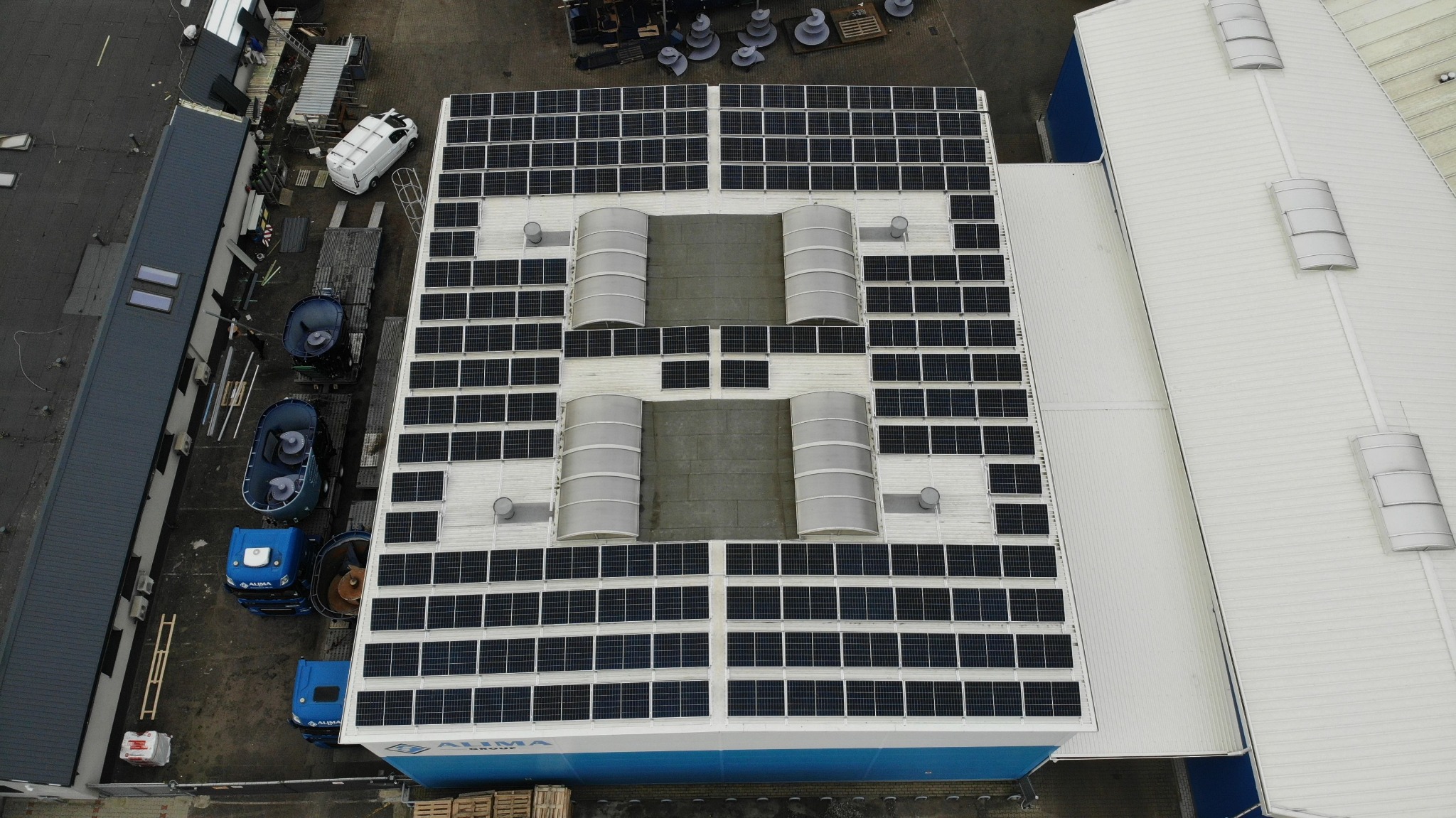 Widok z góry na dach budynku przemysłowego pokryty panelami fotowoltaicznymi z czterema dużymi świetlikami tunelowymi. Panele ułożone w regularną siatkę, z przerwami wokół świetlików. Na dachu...