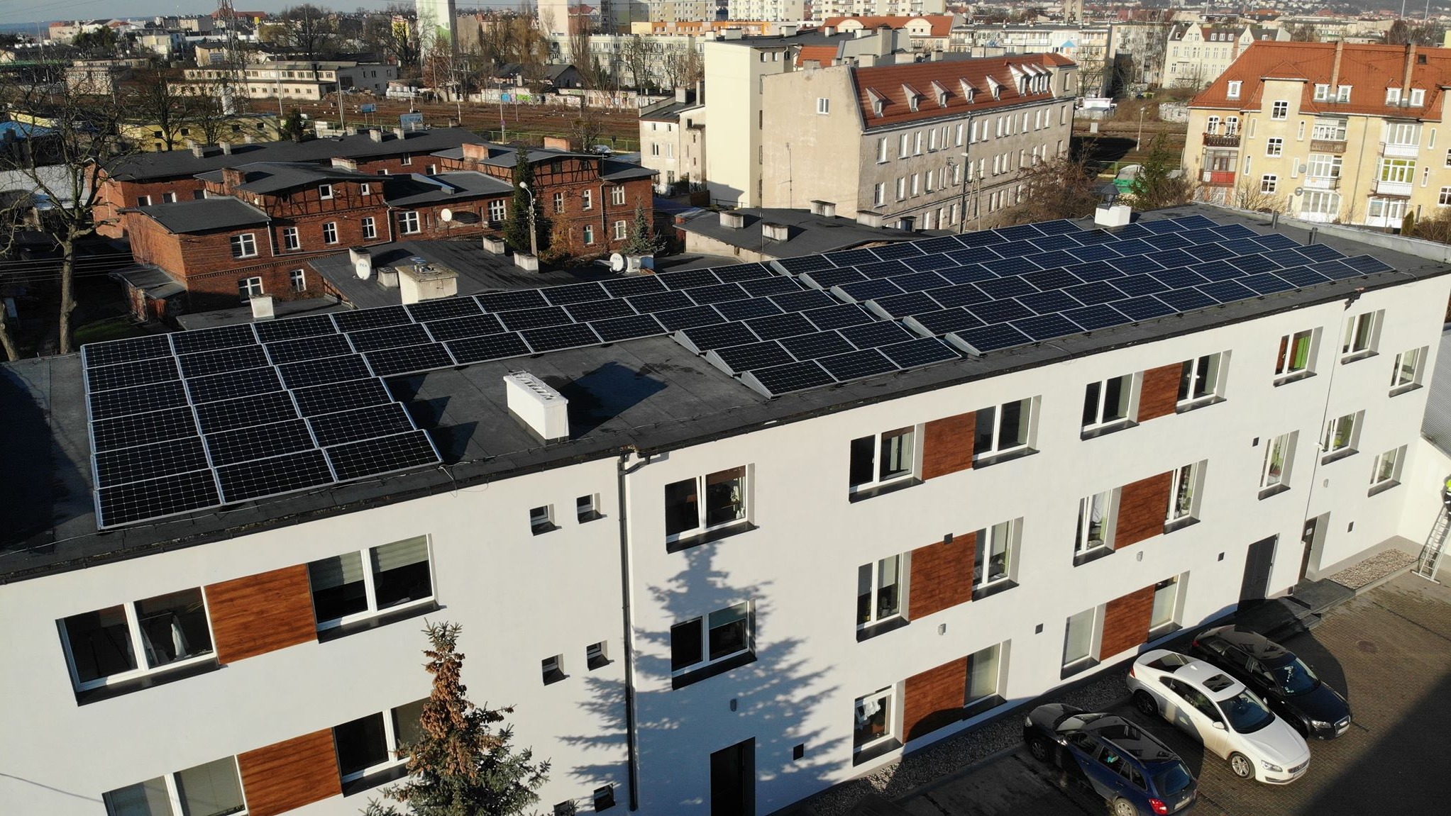Widok z góry na budynek z panelami słonecznymi na dachu, w tle zabudowa miejska, na parkingu zaparkowane trzy samochody, cień drzewa na elewacji.