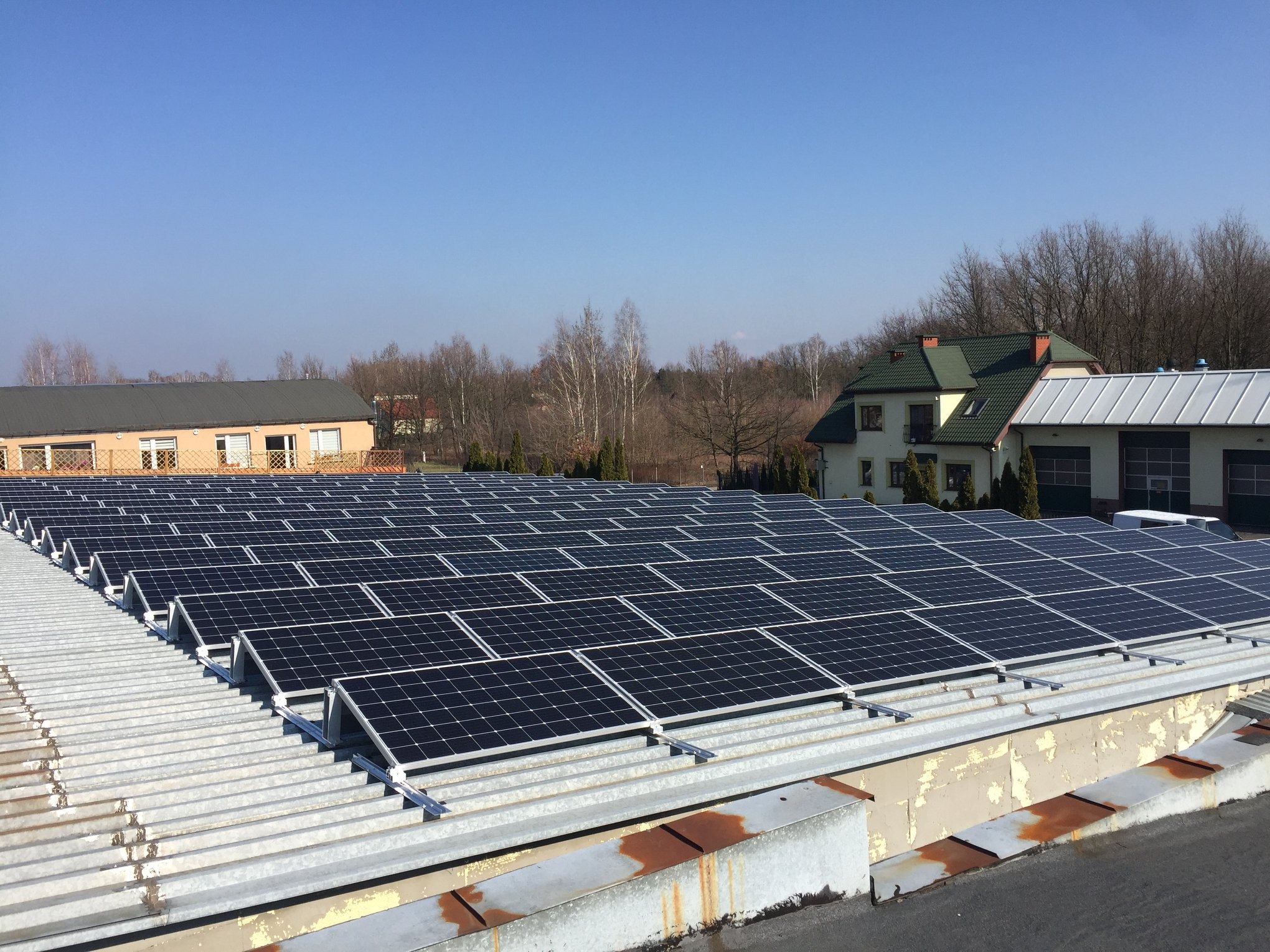 Rząd paneli słonecznych na metalowym dachu budynku przemysłowego w słoneczny dzień, w tle zabudowania i drzewa.