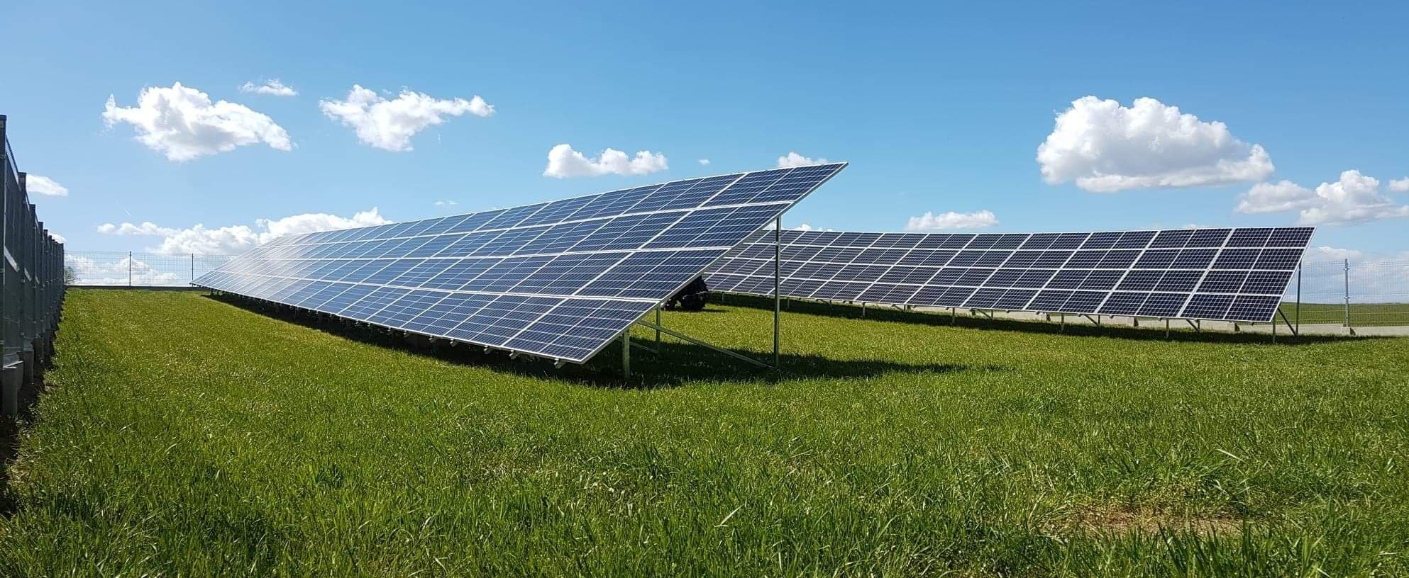 Rozległa farma paneli słonecznych na zielonym polu, pod błękitnym niebem z lekkimi chmurami, widoczne metalowe ogrodzenie w tle.
