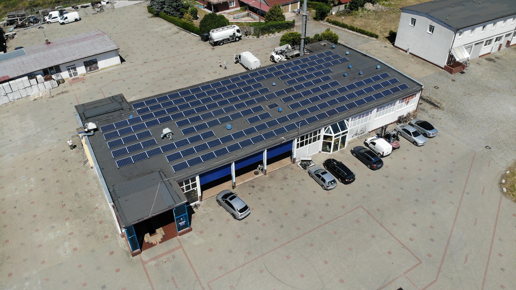 Widok z lotu ptaka na duży budynek z panelami słonecznymi na dachu, zaparkowane samochody i pojazdy dostawcze na placu z kostki brukowej.
