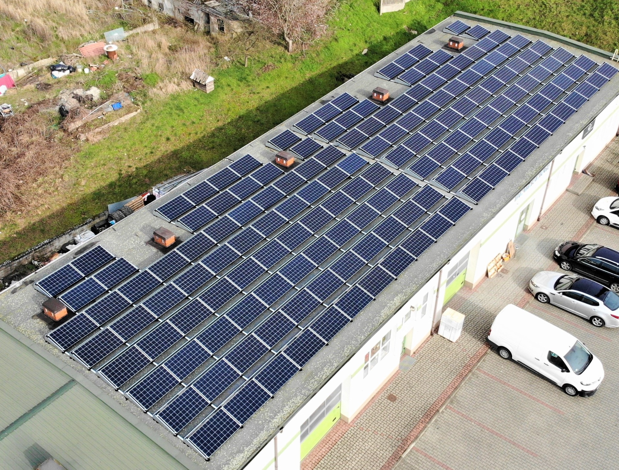 Widok z góry na instalację fotowoltaiczną na dachu budynku w Łomiankach, z widocznymi panelami słonecznymi, elementami wentylacyjnymi i zaparkowanymi samochodami.