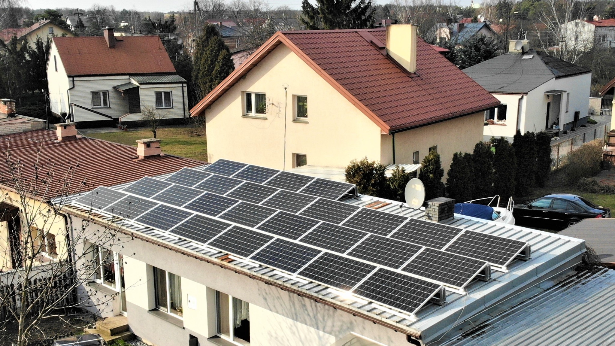 Widok z góry na dom z panelami słonecznymi na dachu, w tle zabudowa jednorodzinna i zieleń, widoczny jacht oraz czarny samochód osobowy.