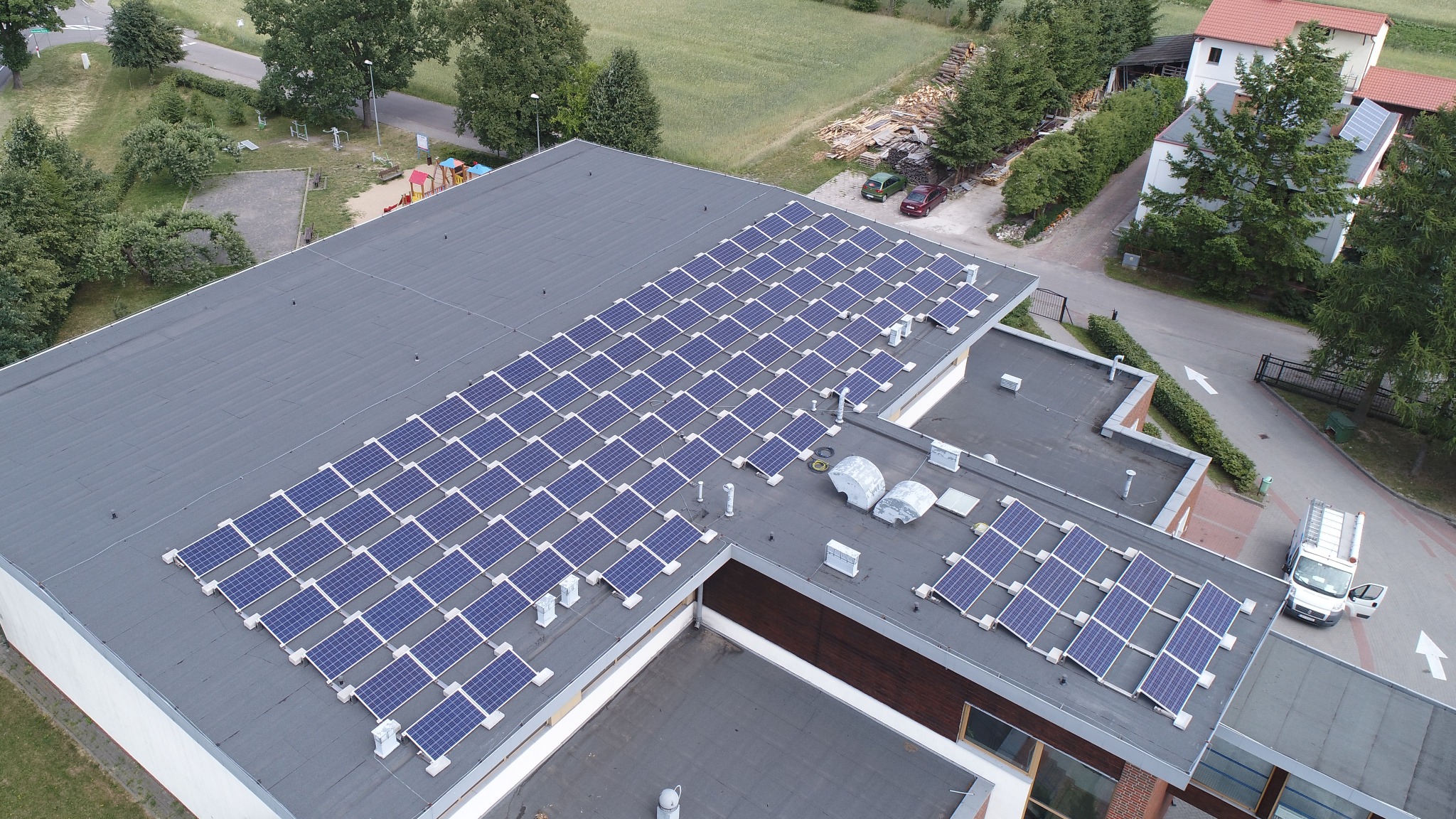 Widok z góry na dużą instalację paneli słonecznych na płaskim dachu budynku, z widocznym placem zabaw i zaparkowanymi samochodami w tle.