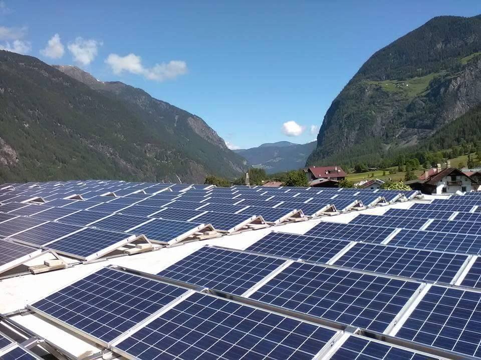 Widok z góry na dużą instalację paneli słonecznych na dachu budynku, z malowniczym krajobrazem górskim w tle i błękitnym niebem z chmurami.