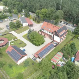 Widok z drona na kompleks budynków z panelami słonecznymi na dachach, otoczonych zielenią i drzewami.