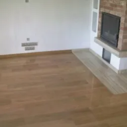 Ułożona podłoga z parkietu w odcieniach brązu, sąsiadująca z wykończeniem kominka z jasnego kamienia.