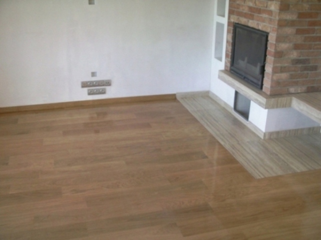 Ułożona podłoga z parkietu w odcieniach brązu, sąsiadująca z wykończeniem kominka z jasnego kamienia.