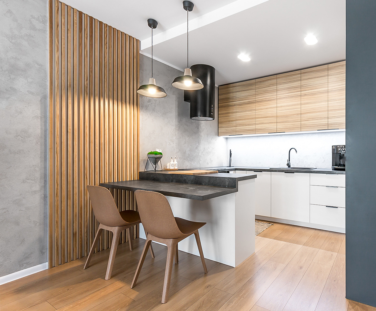 Nowoczesna kuchnia z drewnianymi lamelami na ścianie, szarym blatem i dwoma krzesłami, oświetlona lampami wiszącymi.