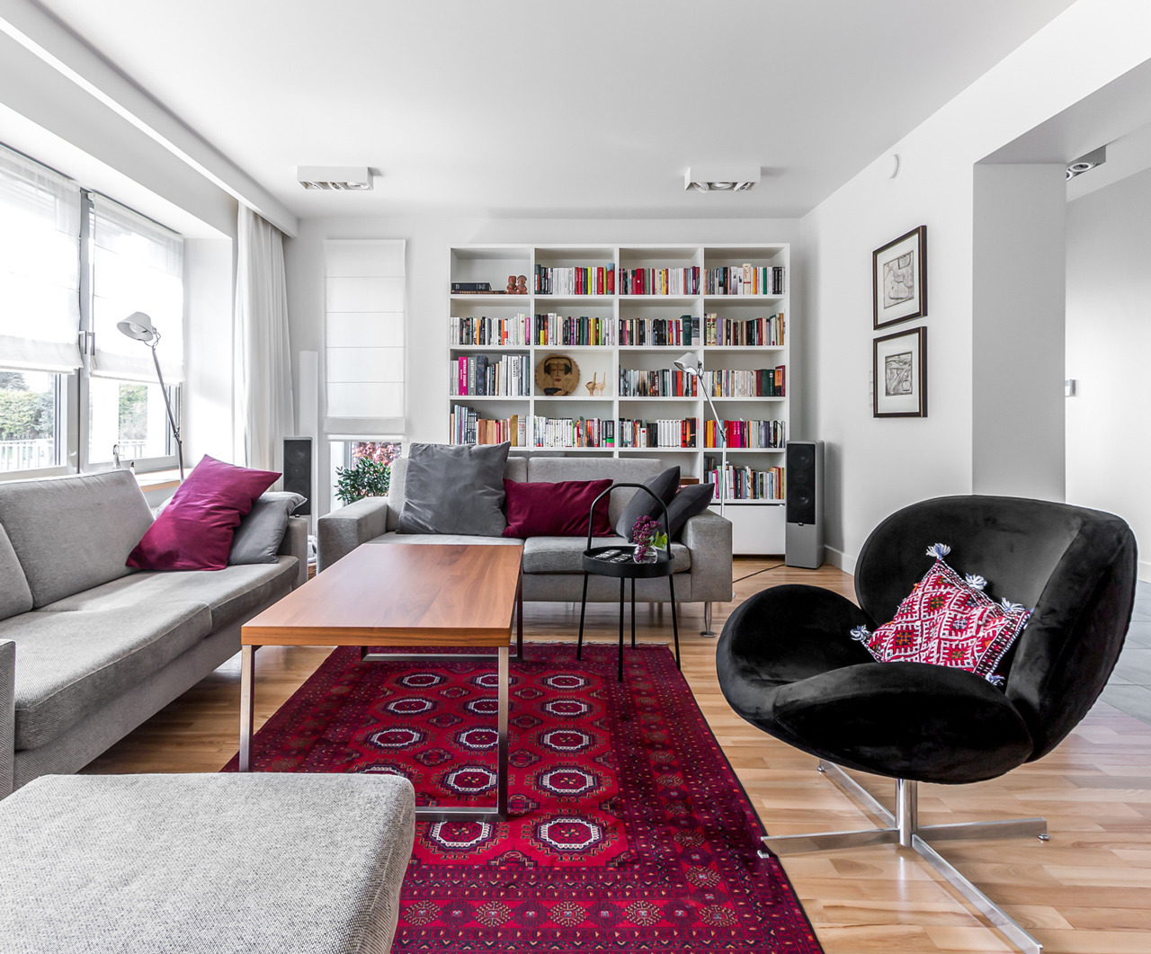 Jasne wnętrze salonu z szarą sofą, drewnianym stolikiem kawowym, wzorzystym czerwonym dywanem i czarnym fotelem typu 'swan' z poduszką w etniczne wzory, na tle białej biblioteczki.