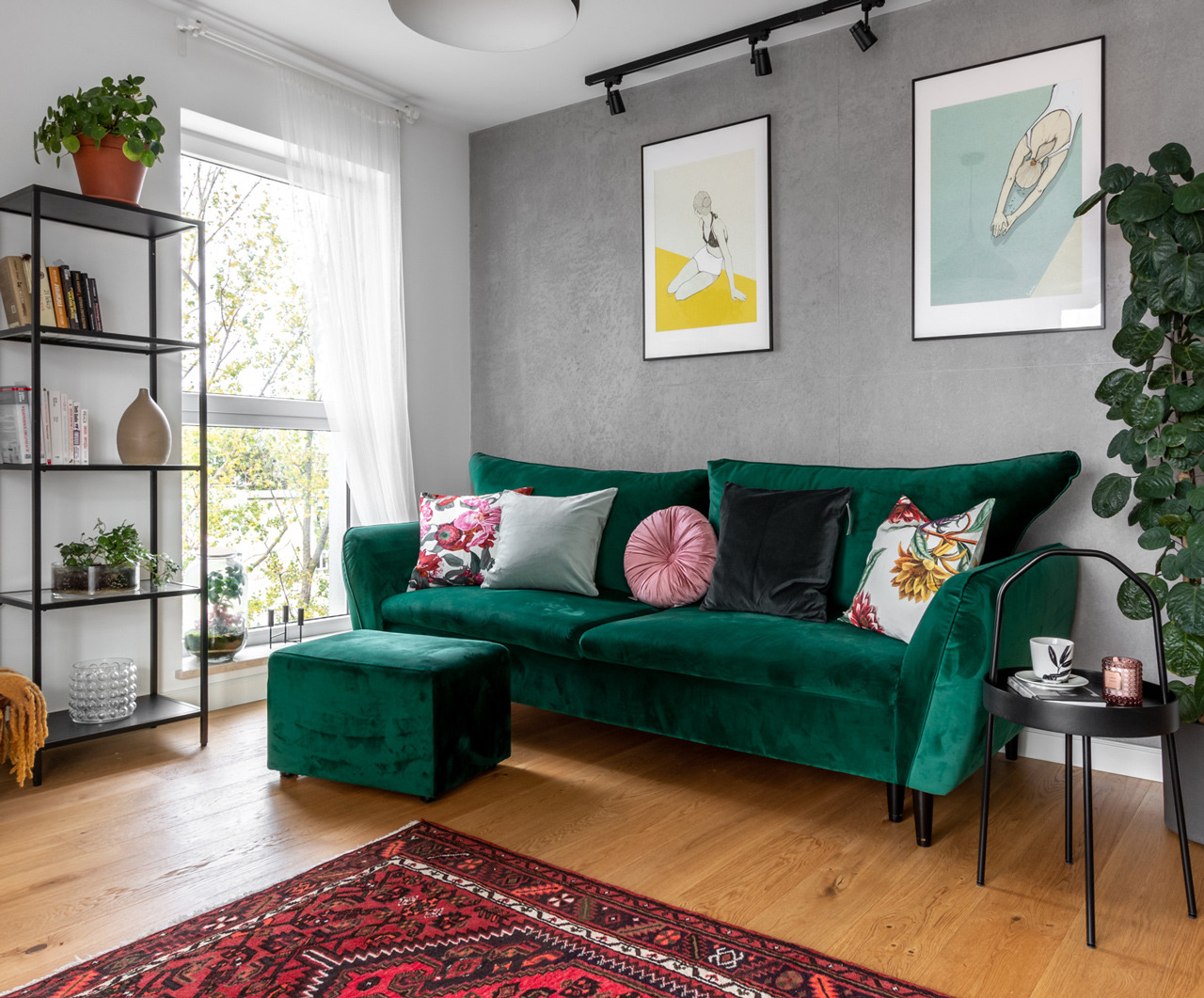Szmaragdowa sofa z poduszkami w salonie, uzupełniona o puf, stolik pomocniczy i regał z dekoracjami, na drewnianej podłodze z orientalnym dywanem.