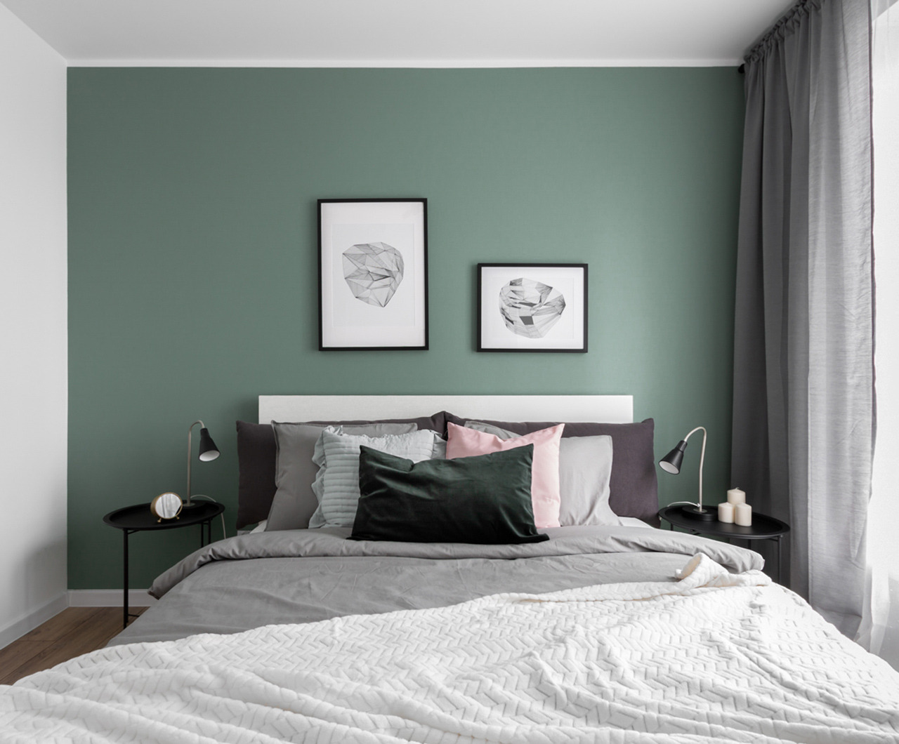Jasne wnętrze sypialni z łóżkiem, zieloną ścianą, minimalistycznymi grafikami w czarnych ramkach i szarą zasłoną przy oknie.