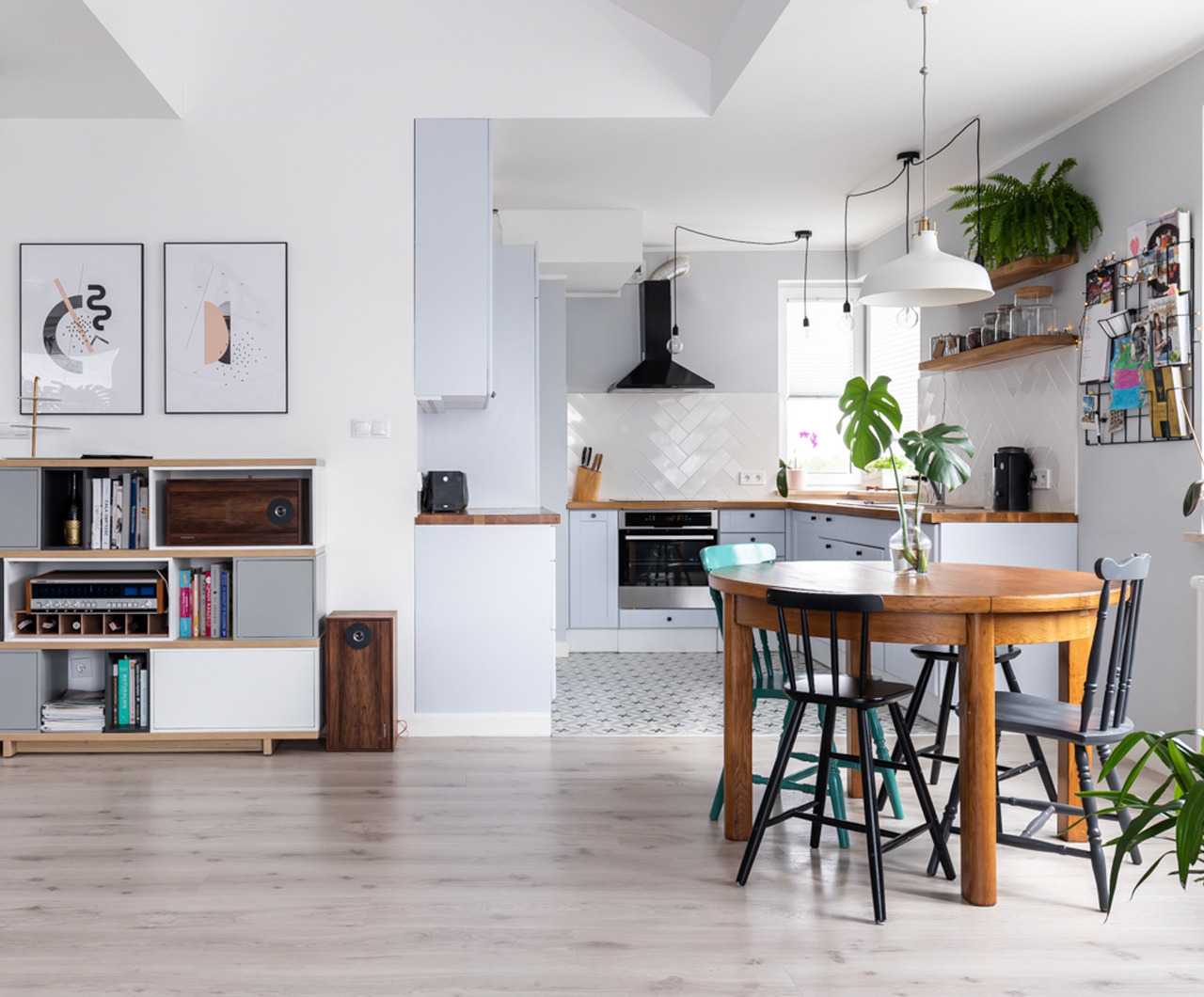Jasne wnętrze mieszkania z otwartą kuchnią, drewniany stół z krzesłami, regał z książkami i dekoracjami, biało-szara kolorystyka, podłoga z paneli, nowoczesne oświetlenie wiszące.