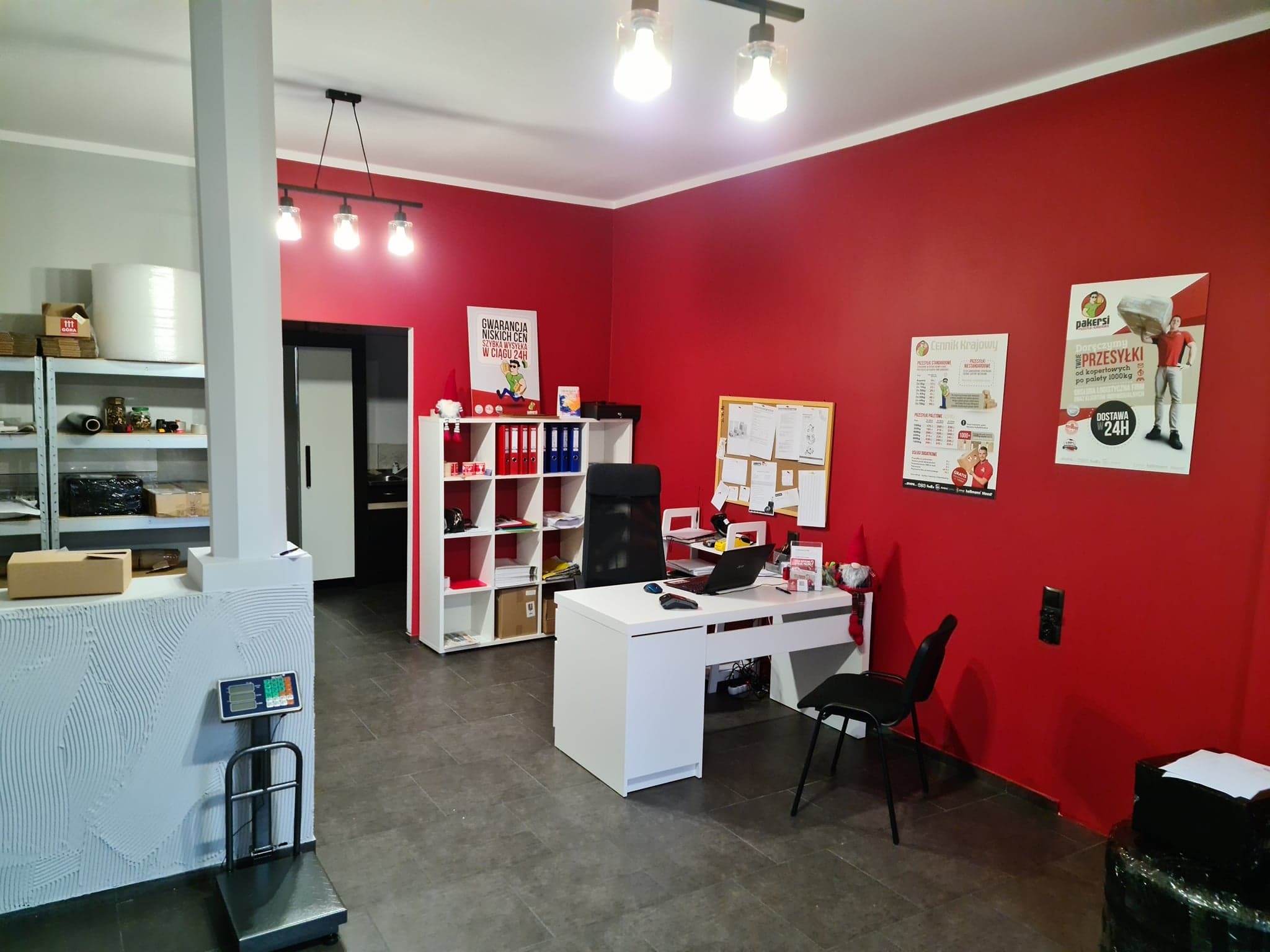 Wnętrze biura firmy transportowej z wagą platformową, biurkiem, regałami i plakatami reklamowymi na czerwonej ścianie.