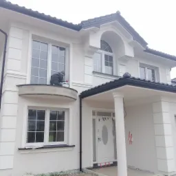 Pracownik w szarej kurtce maluje elewację dwupiętrowego, białego domu z czarnym dachem, siedząc na balkonie i trzymając białe wiadro z farbą.