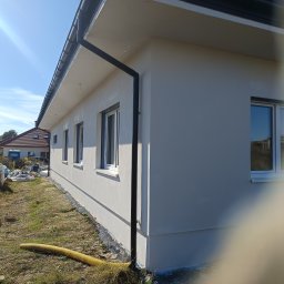 ARIA Kamil Kasprzak - Ściana budynku po tynkowaniu, widoczne okna i rynna spustowa. W tle gruz i materiały budowlane. Słoneczny dzień, błękitne niebo.