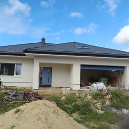 Kamil Kasprzak - Nowoczesny dom jednorodzinny z szarą dachówką, jasną elewacją i niebieskimi drzwiami. Widoczne rusztowania i materiały budowlane. Błękitne niebo z chmurami w tle.