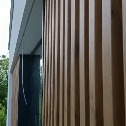 Elewacja budynku z drewnianymi lamelami, widok z dołu, jasne drewno, nowoczesna architektura, okno z czarną ramą, fragment zieleni w tle.