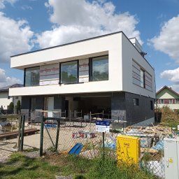 Kamil Kasprzak - Nowoczesny dom w trakcie elewacji: biały tynk na górze, ciemny kamień na dole. Okna zabezpieczone folią, widoczny pracownik na dachu. Żółta skrzynka gazowa na pierwszym planie.