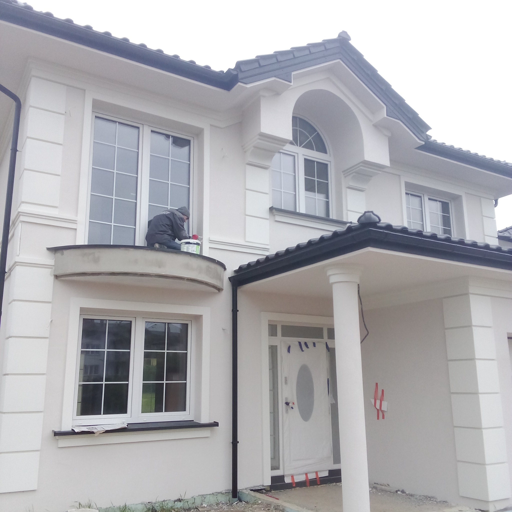 Pracownik w szarej kurtce maluje elewację dwupiętrowego, białego domu z czarnym dachem, siedząc na balkonie i trzymając białe wiadro z farbą.