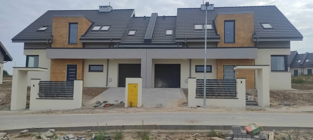 Nowoczesny bliźniak z szarą dachówką, jasną elewacją z drewnianymi akcentami i antracytowymi oknami, widok z ulicy, w tle kolejne domy w zabudowie szeregowej.