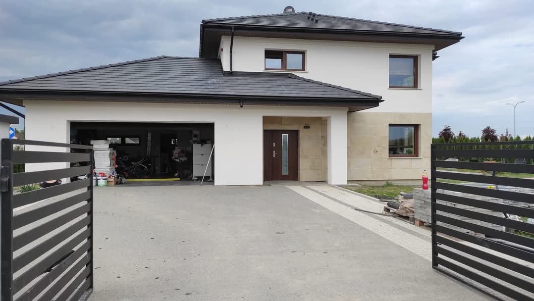 Nowoczesny dom jednorodzinny z garażem, jasną elewacją i elementami z kamienia naturalnego, widok zza otwartej, metalowej bramy wjazdowej, materiały budowlane składowane obok.