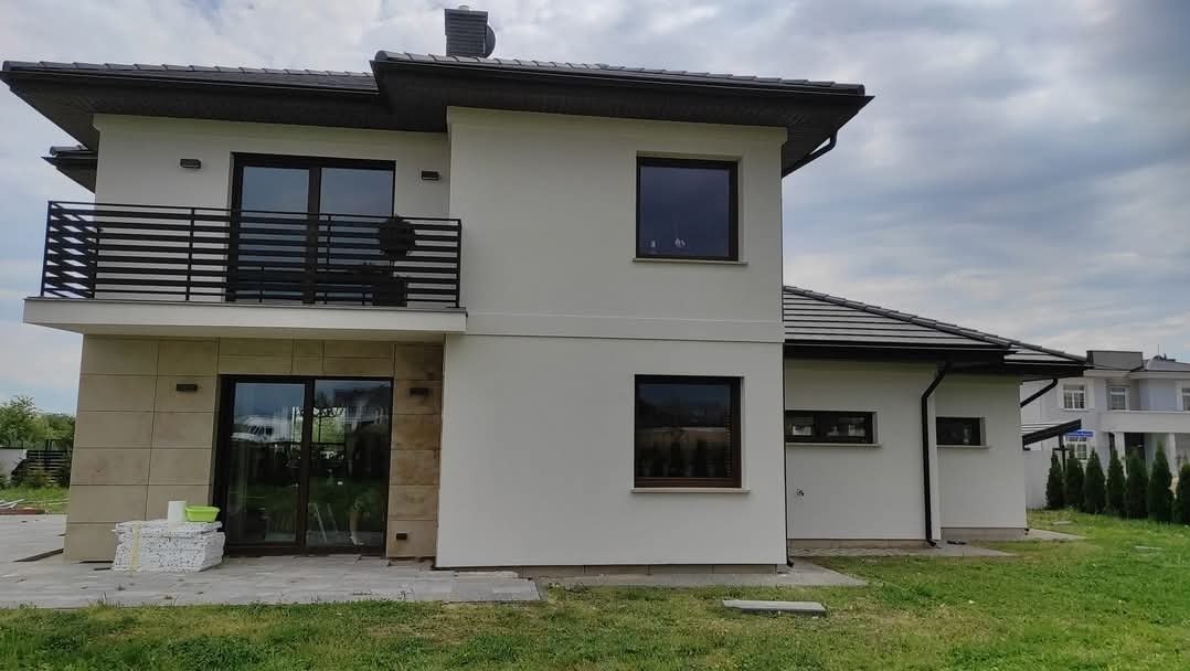 Nowoczesny dom jednorodzinny z białymi ścianami i balkonem z czarną balustradą, widoczne materiały izolacyjne przy drzwiach tarasowych.