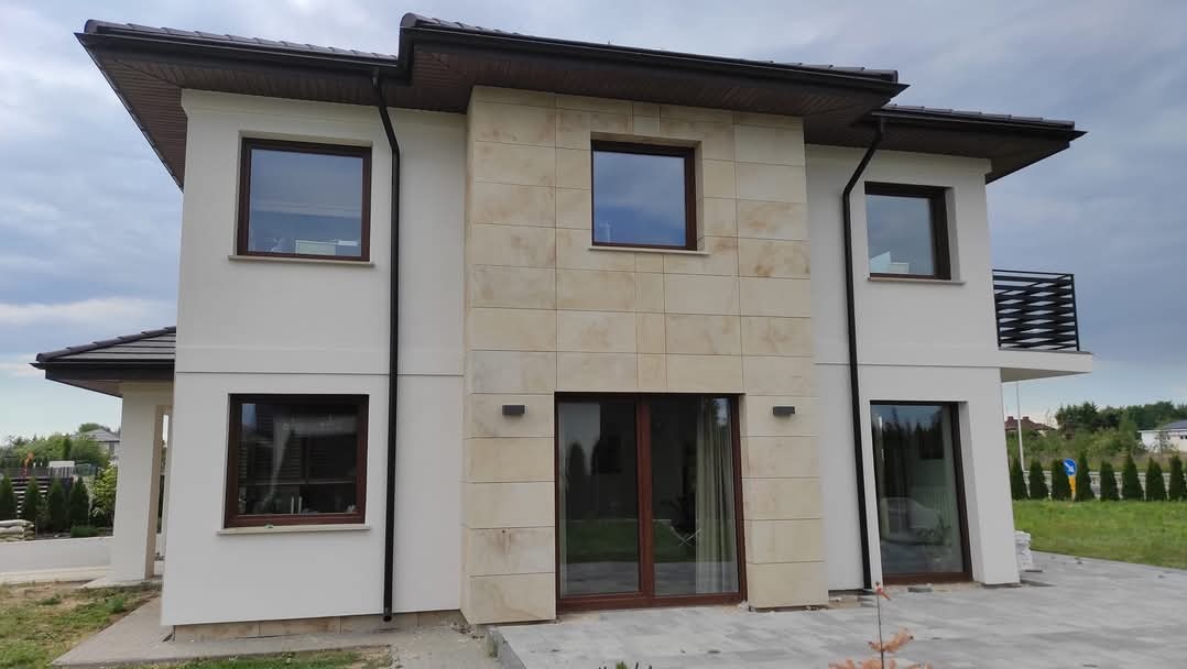 Nowoczesny dom jednorodzinny w Lublinie z elewacją częściowo wykończoną beżowymi płytami kamiennymi, widoczne okna z brązowymi ramami i ciemny dach.