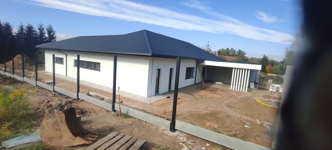 Nowoczesny, parterowy dom z białymi ścianami i ciemną dachówką, widoczny na etapie wykończenia terenu wokół budynku, z fragmentem koparki w kadrze.