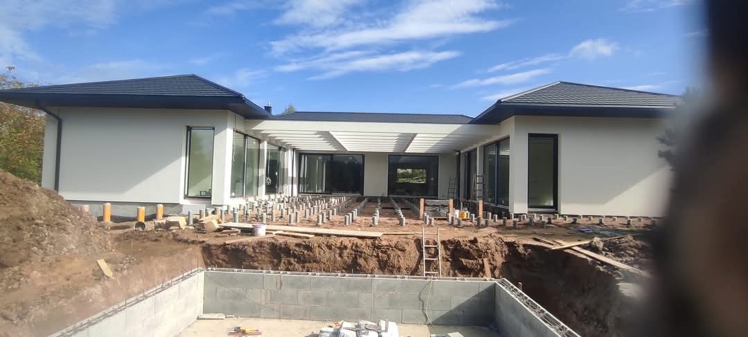 Nowoczesny dom jednorodzinny z jasną elewacją w trakcie budowy tarasu, widoczne fundamenty basenu i przygotowane podłoże pod deski tarasowe.