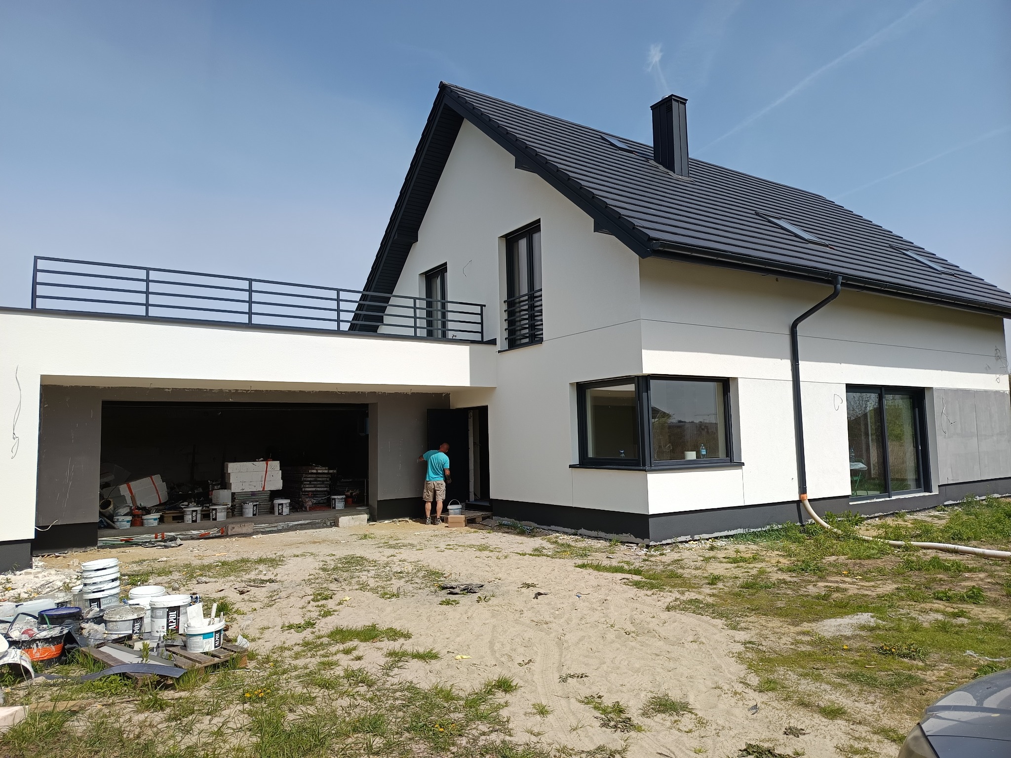 Nowoczesny dom z białą elewacją w trakcie wykończenia. Widoczny pracownik przy wejściu i puste wiadra po farbie. Część elewacji jeszcze nieotynkowana.