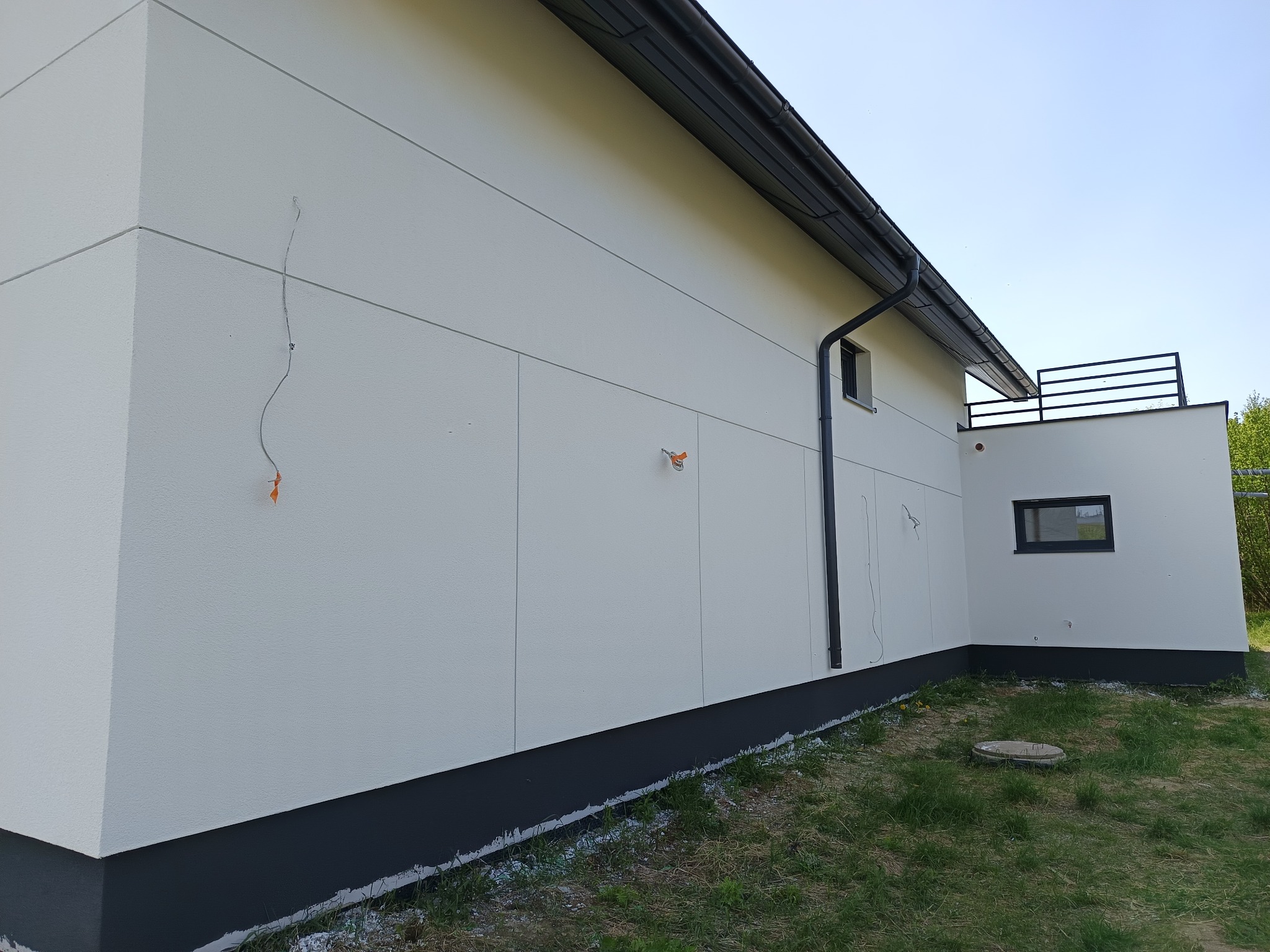 Nowoczesna elewacja budynku w jasnych odcieniach szarości z ciemną podmurówką. Widoczne fragmenty instalacji elektrycznej i okno z czarną ramą.