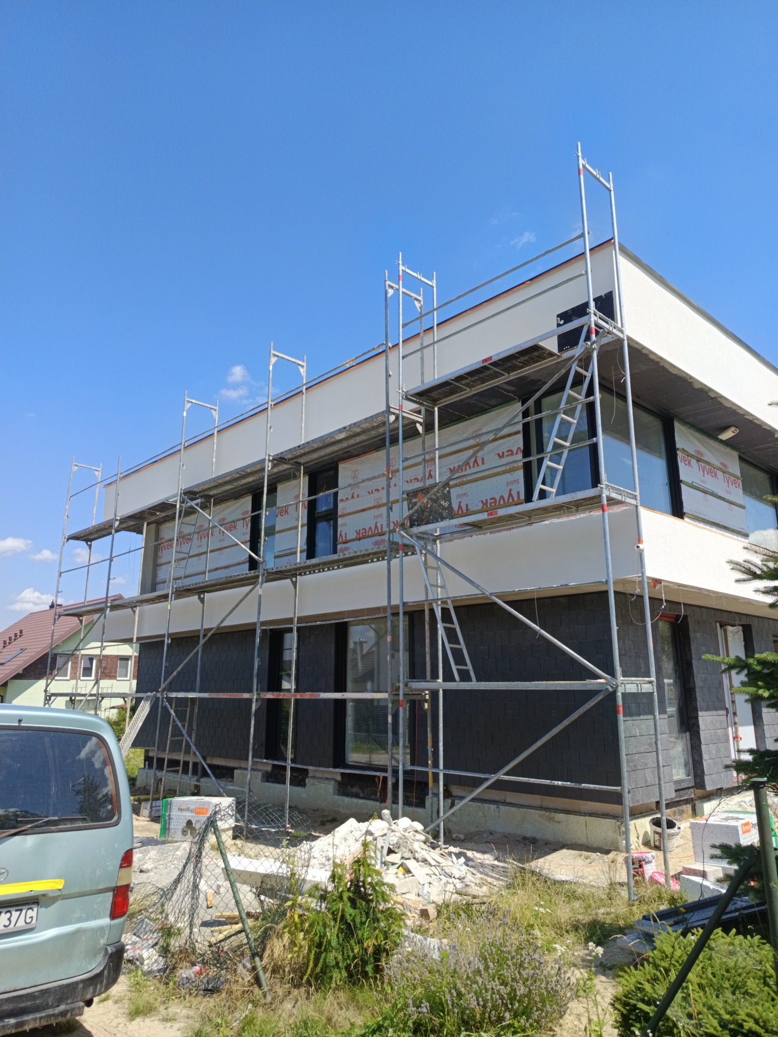 Dom w trakcie elewacji: rusztowania, folia ochronna Tyvek, częściowo otynkowana ściana i wykończona szarą okładziną. Błękitne niebo w tle.