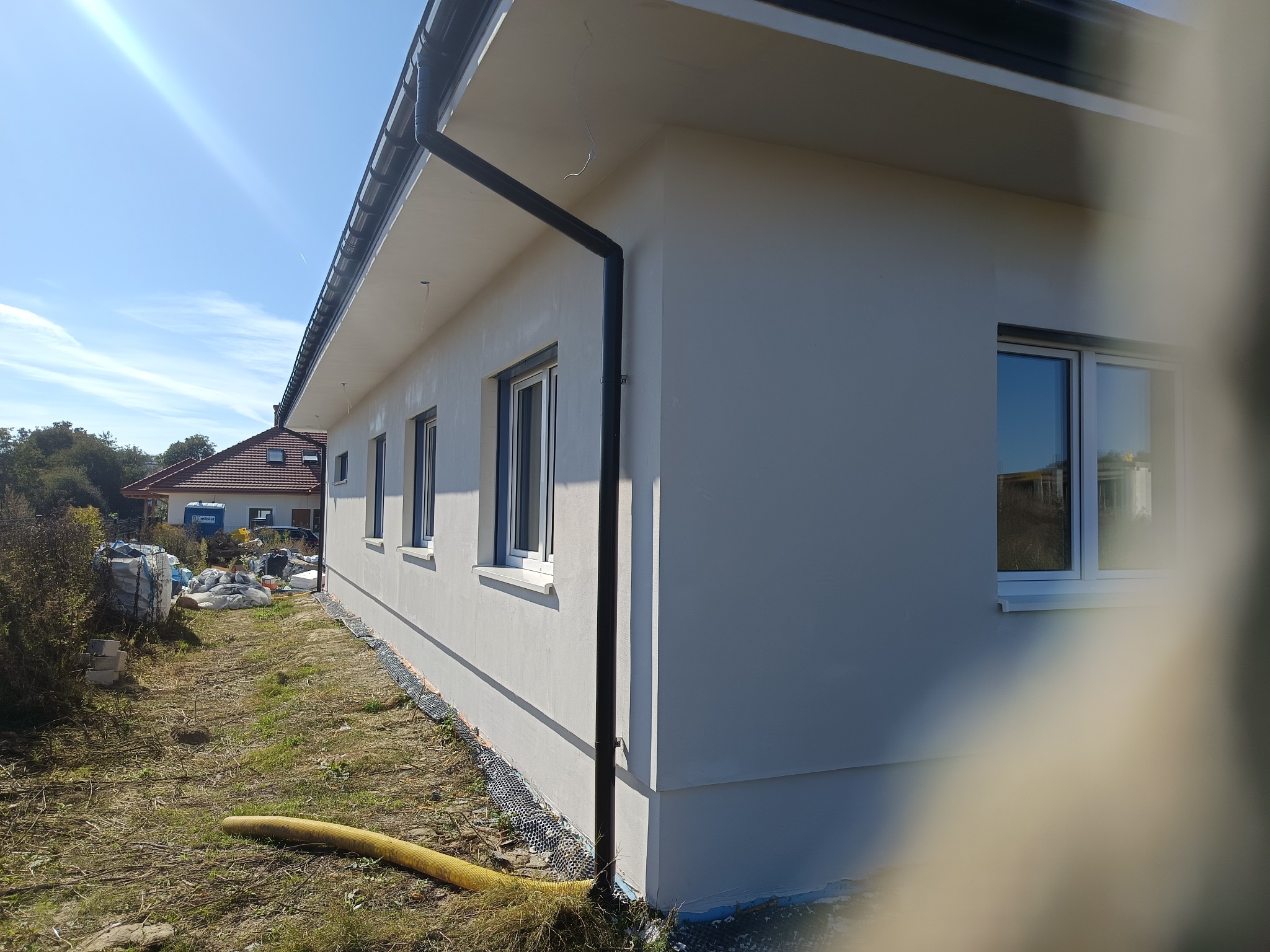 Ściana budynku po tynkowaniu, widoczne okna i rynna spustowa. W tle gruz i materiały budowlane. Słoneczny dzień, błękitne niebo.