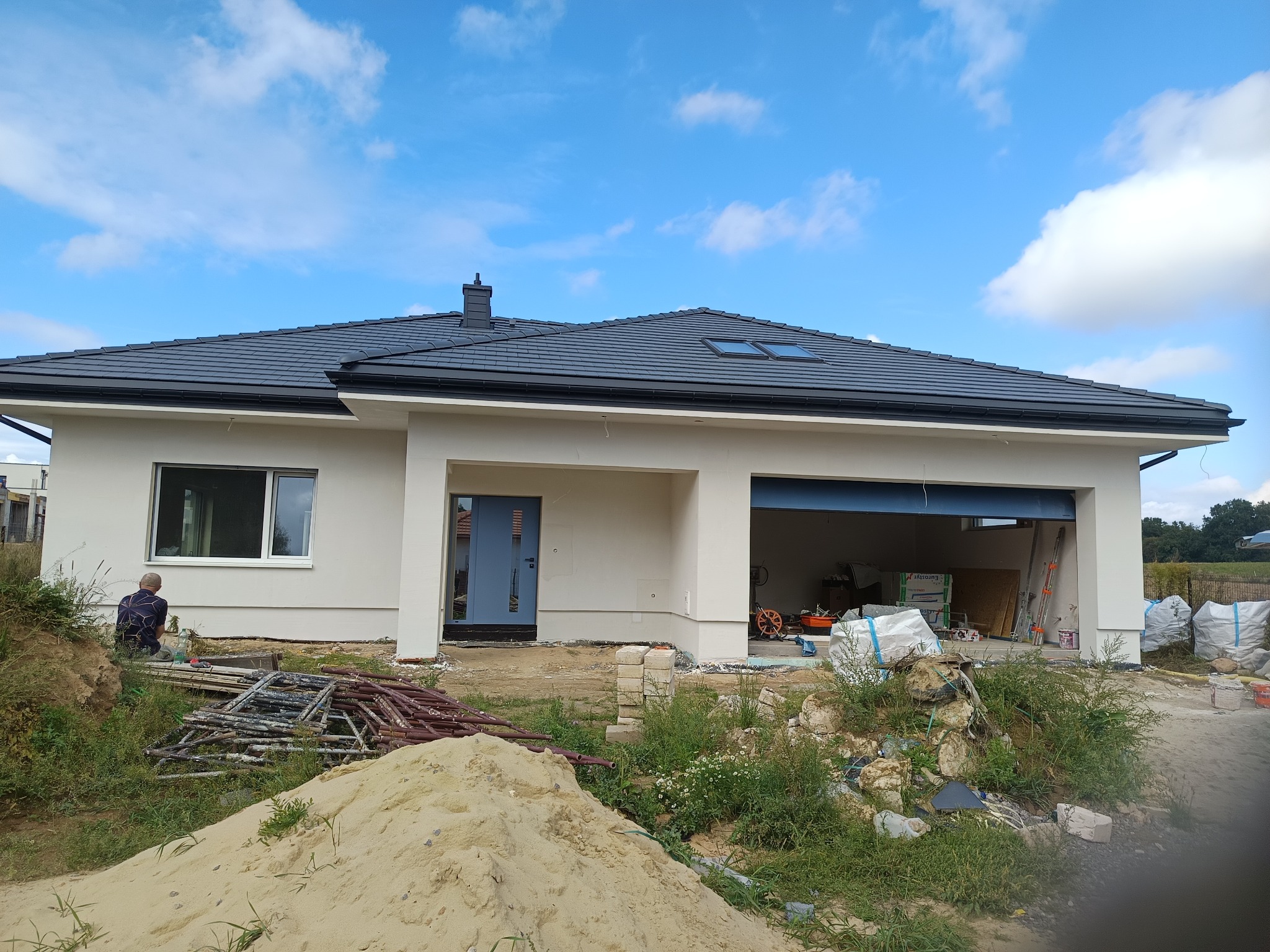 Nowoczesny dom jednorodzinny z szarą dachówką, jasną elewacją i niebieskimi drzwiami. Widoczne rusztowania i materiały budowlane. Błękitne niebo z chmurami w tle.