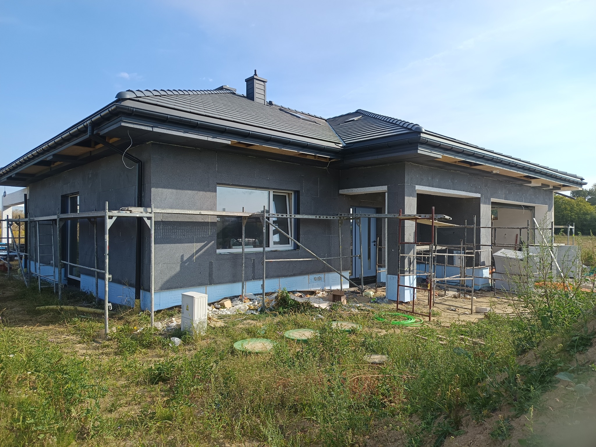 Surowy dom jednorodzinny w Lublinie, w trakcie ocieplania, z widocznymi rusztowaniami i nieotynkowaną elewacją. Budynek z szarym dachem, okna i drzwi zamontowane.