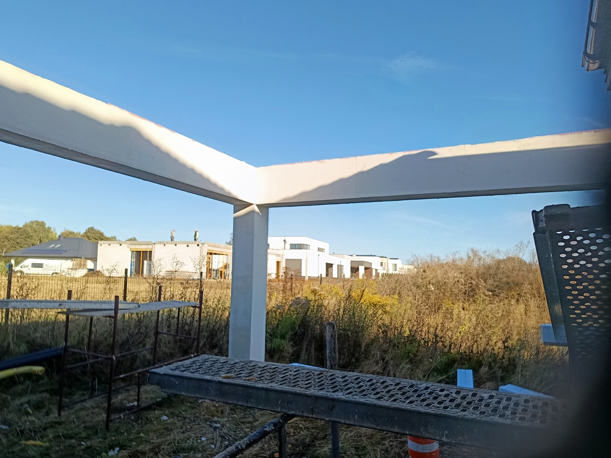 Surowy szkielet domu w budowie: betonowe słupy i belki na tle krajobrazu. Widoczne rusztowania i elementy konstrukcyjne. Ujęcie z dołu, błękitne niebo w tle.