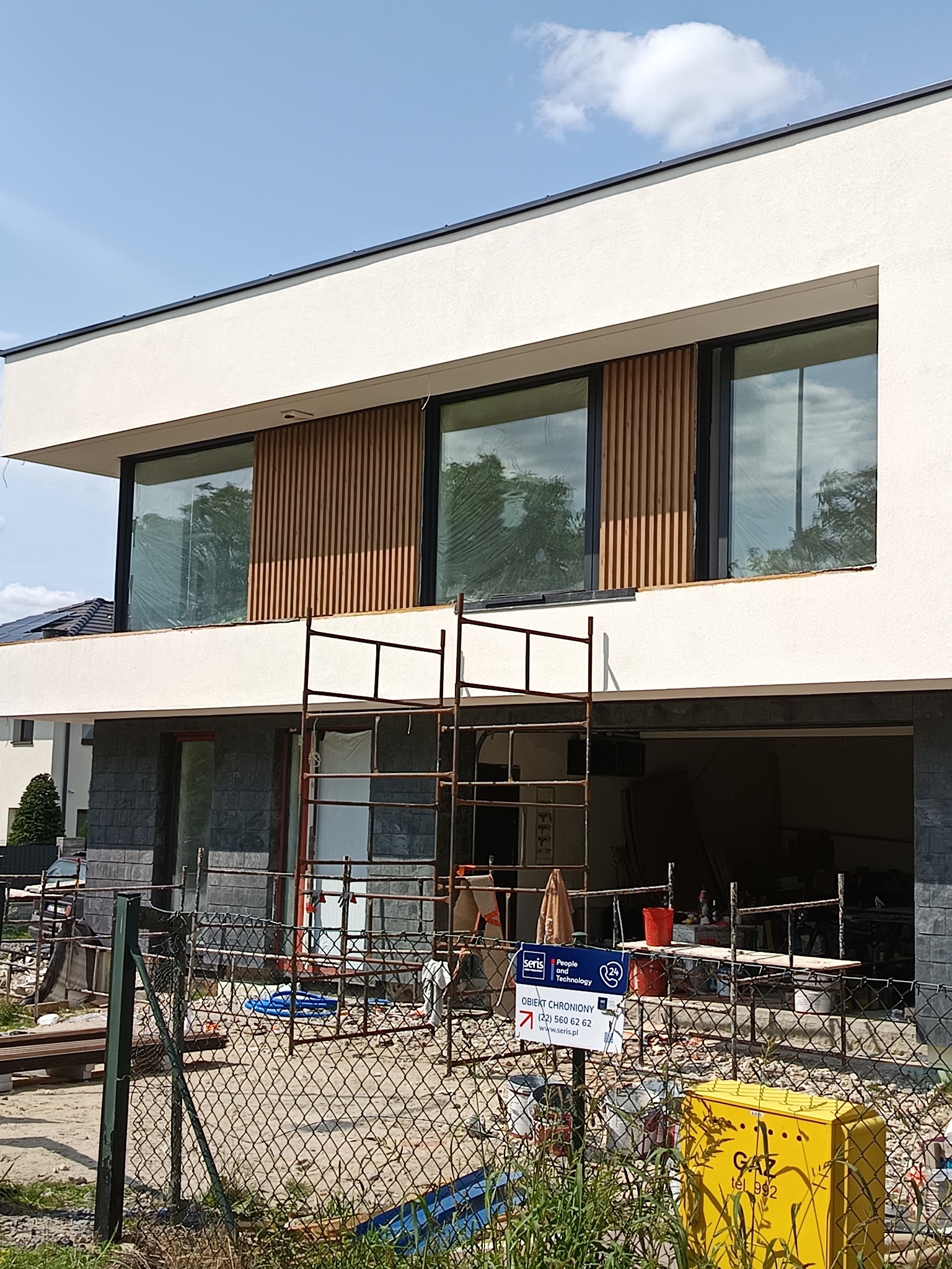 Nowoczesny dom w trakcie wykończenia elewacji, widoczne rusztowania. Biały tynk kontrastuje z drewnianymi panelami między oknami. Dolna część budynku wykończona szarą cegłą.