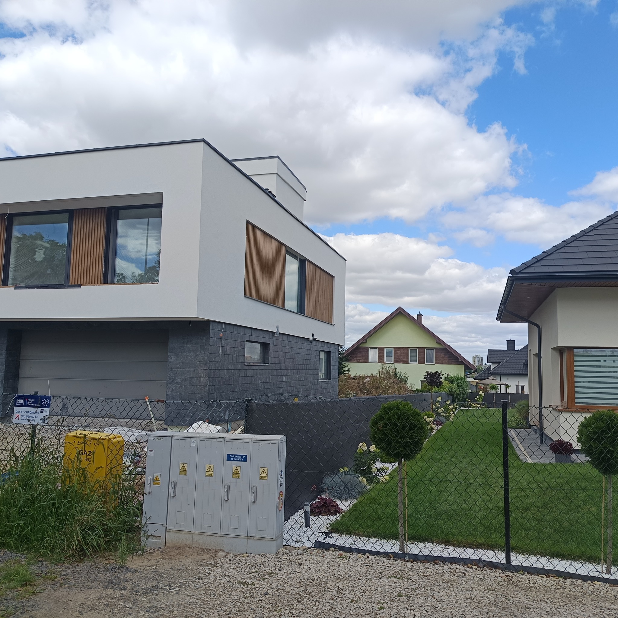 Nowoczesny dom z elewacją w bieli i szarości, drewniane żaluzje okienne, zielony trawnik za ogrodzeniem z siatki. Błękitne niebo z obłokami w tle.