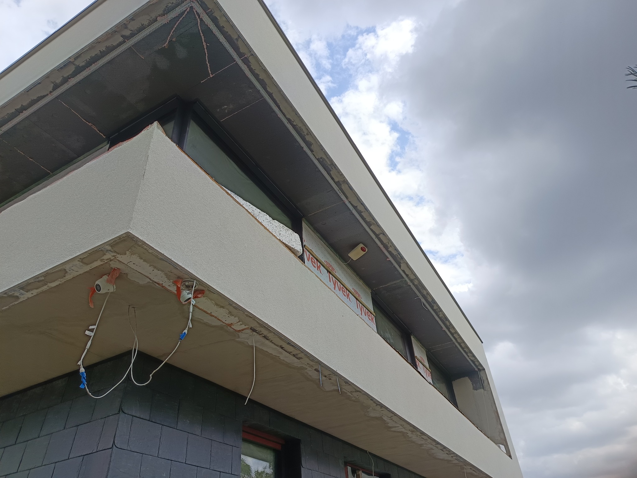 Fragment elewacji budynku w trakcie prac wykończeniowych; widoczne ocieplenie, tynk i instalacje elektryczne. Ujęcie z dołu na tle pochmurnego nieba.