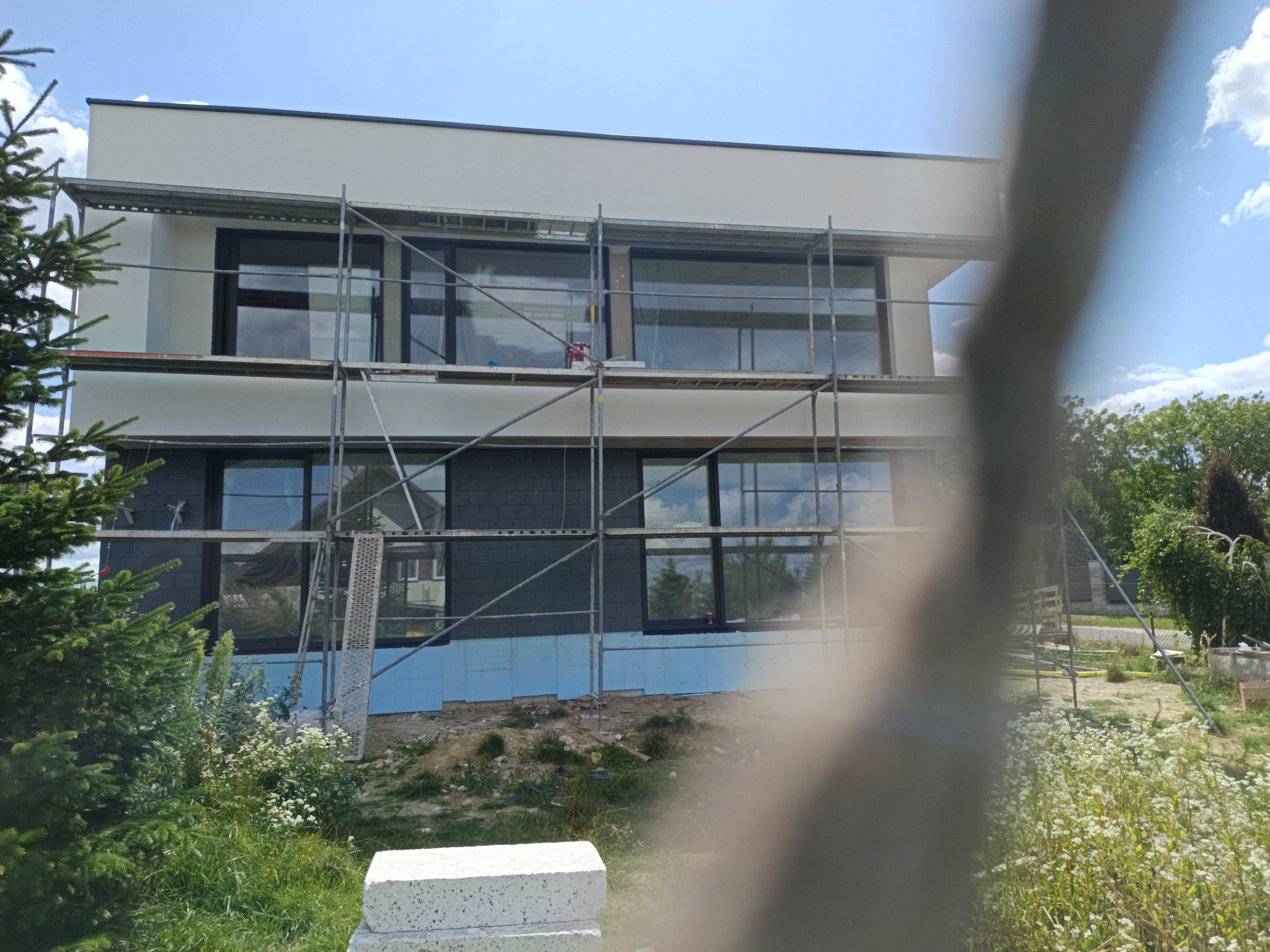 Dwukondygnacyjny dom w trakcie ocieplania, z widocznymi rusztowaniami i izolacją termiczną na elewacji, otoczony zielenią i elementami budowlanymi.