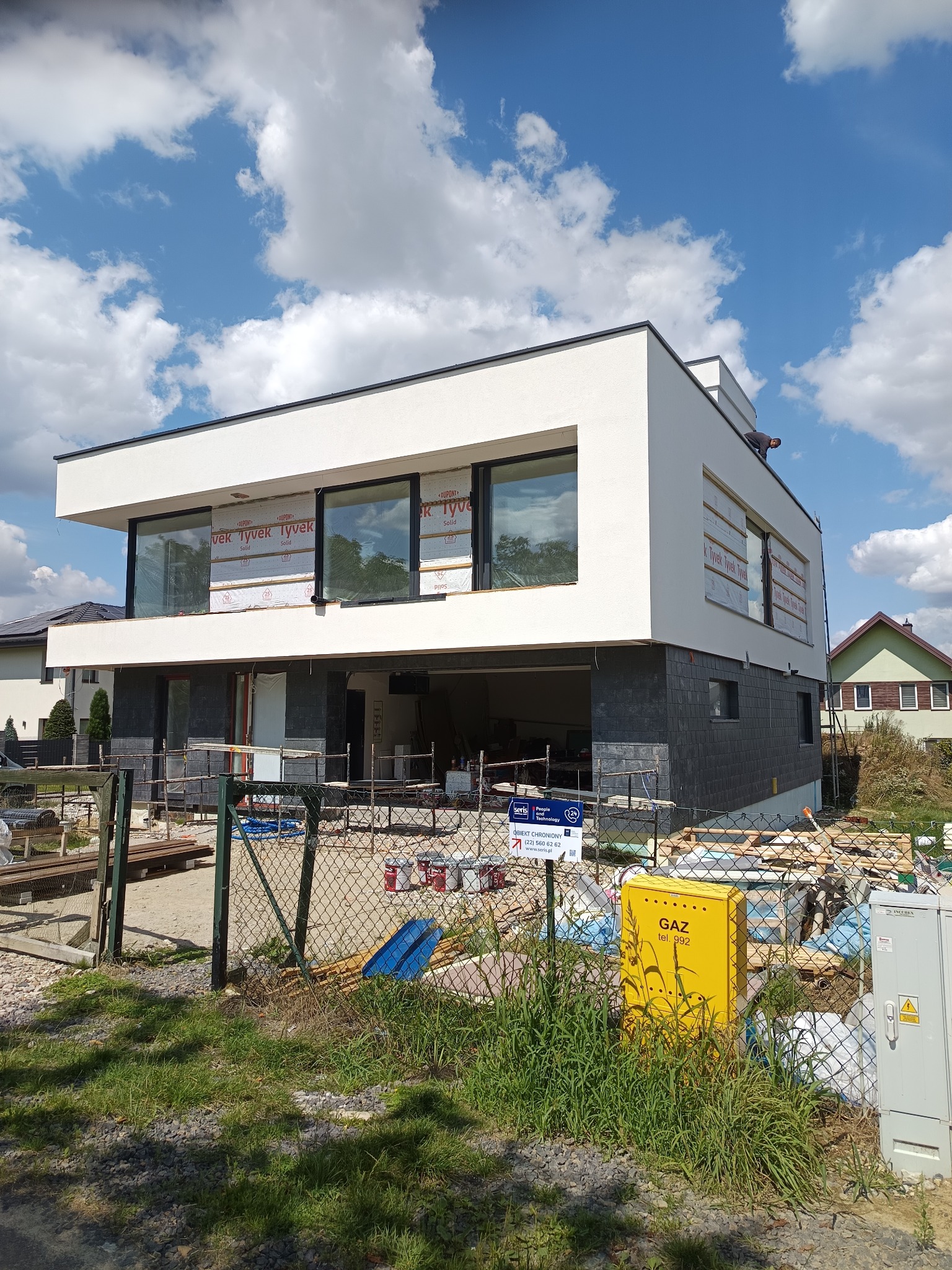 Nowoczesny dom w trakcie elewacji: biały tynk na górze, ciemny kamień na dole. Okna zabezpieczone folią, widoczny pracownik na dachu. Żółta skrzynka gazowa na pierwszym planie.