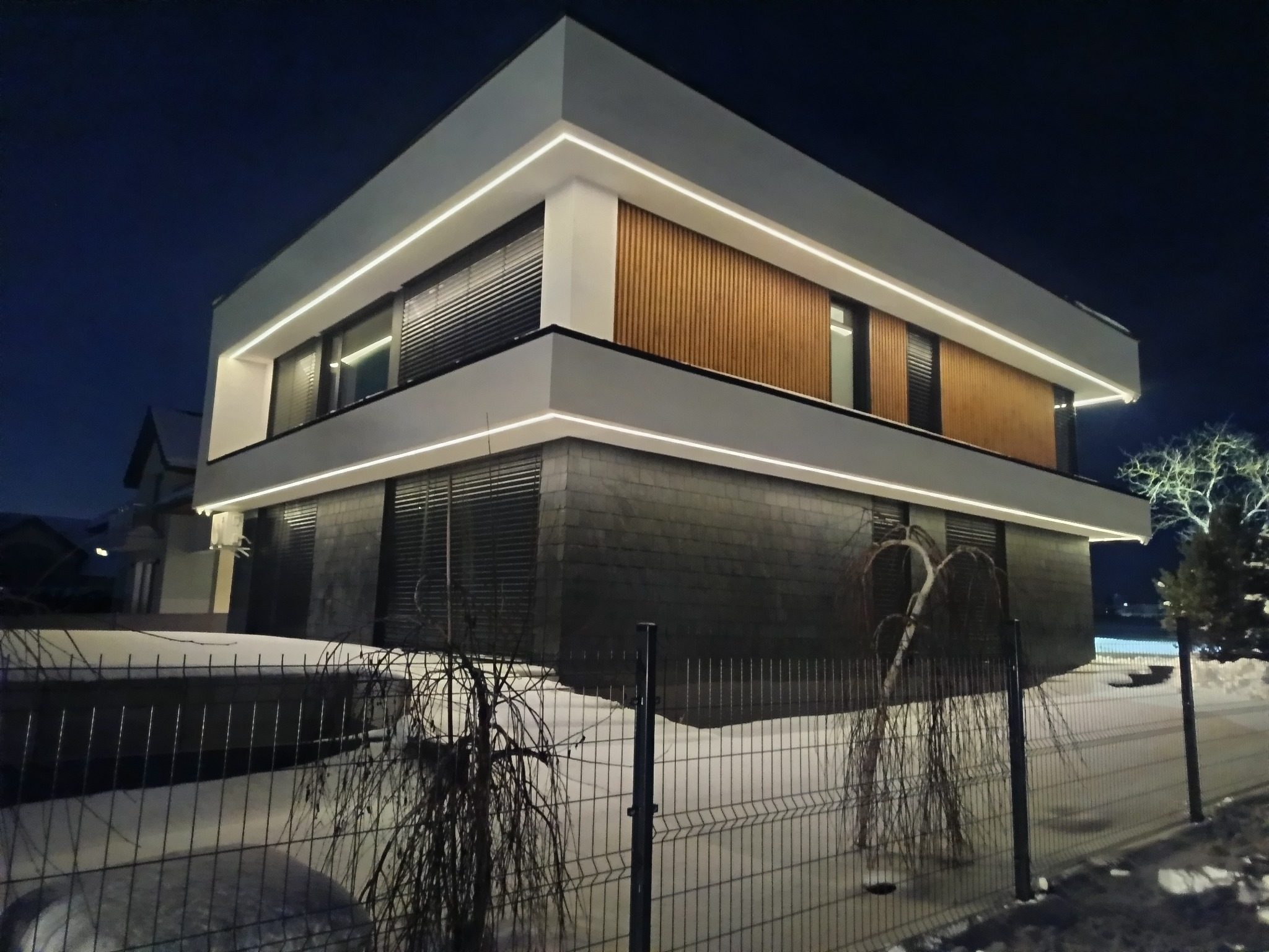 Nowoczesny dom z podświetloną elewacją w nocy. Widoczne drewniane panele na piętrze i ciemna okładzina na parterze. Świecące linie LED podkreślają architekturę budynku.