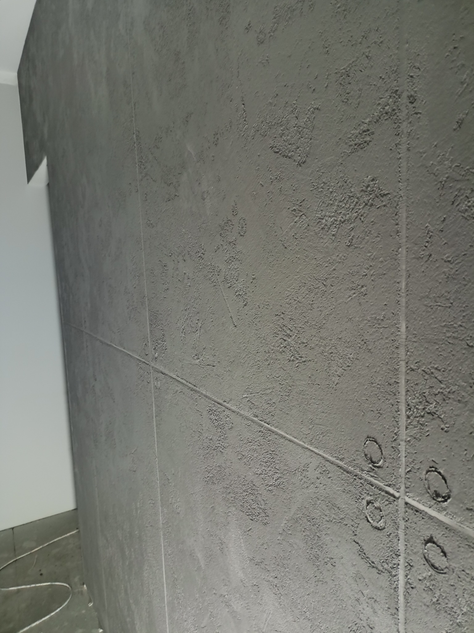 Szara ściana z fakturą, podzielona pionowymi i poziomymi liniami, z widocznymi otworami na gniazdka elektryczne, w trakcie prac wykończeniowych.